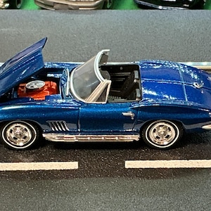 May include: A blue convertible toy car with the hood open, revealing a red engine. The car has chrome accents and white-walled tires. The car is on a black surface with white lines.