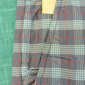 May include: Close-up of a blue, white, and red plaid fabric with a pocket. A yellow rectangular object is partially visible in the pocket. The fabric is a mix of light and dark blue, white, and red threads.