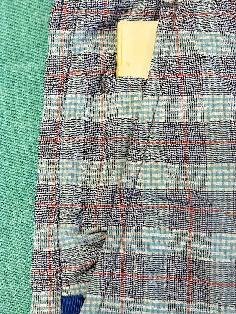 May include: Close-up of a blue, white, and red plaid fabric with a pocket. A yellow rectangular object is partially visible in the pocket. The fabric is a mix of light and dark blue, white, and red threads.