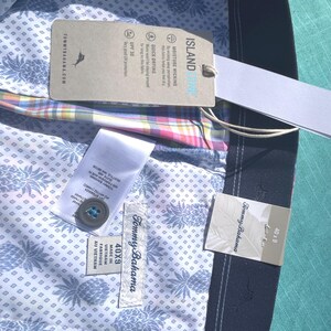 K&ouml;nnte beinhalten: Nahaufnahme eines Tommy Bahama Hemdes mit Etiketten. Das Hemd hat ein hellblaues Ananasmuster auf wei&szlig;em Hintergrund. Die Etiketten enthalten "ISLAND", "MOISTURE WICKING" und "QUICK DRYING". Die Gr&ouml;&szlig;e ist 40x9.
