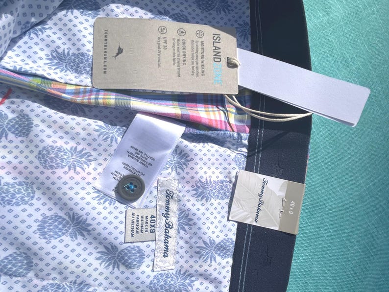 K&ouml;nnte beinhalten: Nahaufnahme eines Tommy Bahama Hemdes mit Etiketten. Das Hemd hat ein hellblaues Ananasmuster auf wei&szlig;em Hintergrund. Die Etiketten enthalten "ISLAND", "MOISTURE WICKING" und "QUICK DRYING". Die Gr&ouml;&szlig;e ist 40x9.