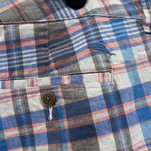 Peut inclure: Gros plan sur un short &agrave; carreaux dans les tons de bleu, rose, blanc et gris. Le short a une fermeture boutonn&eacute;e et une poche. Un petit poisson brod&eacute; bleu est visible sur le short.