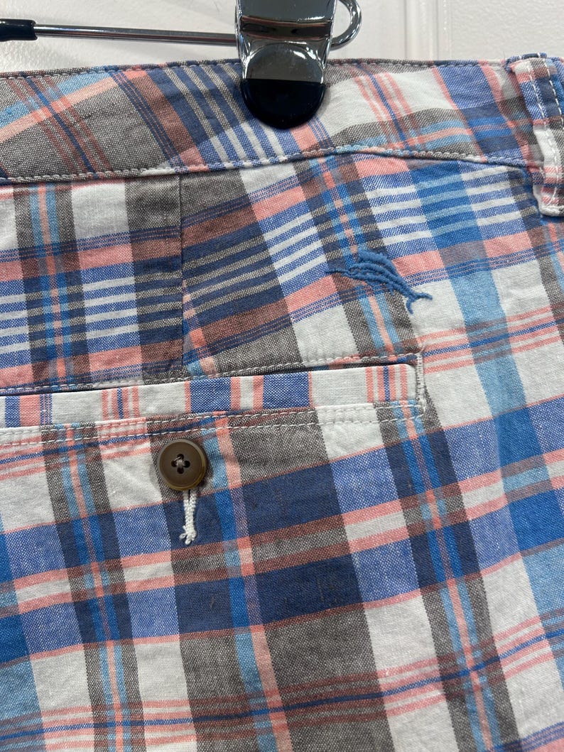 Peut inclure: Gros plan sur un short &agrave; carreaux dans les tons de bleu, rose, blanc et gris. Le short a une fermeture boutonn&eacute;e et une poche. Un petit poisson brod&eacute; bleu est visible sur le short.