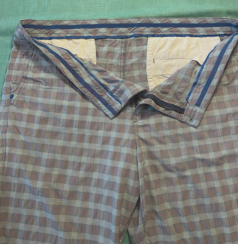 May include: A pair of plaid shorts in shades of gray, blue, and white. The shorts have a button and zipper closure, with pockets visible. The waistband features blue stripes. The fabric has a checkered pattern.