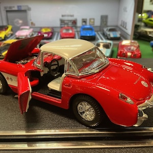 May include: A red and white toy car with the hood and door open, showcasing interior details. The car has chrome accents and sits on a black track. Other toy cars are visible in the background.
