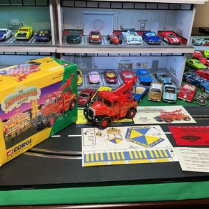 May include: A collection of toy cars and a box set on a black surface. The box is yellow and green with the text "The Showman's Range" and a red tow truck illustration. A red tow truck model and other toy cars are also visible.