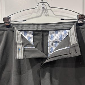 May include: Gray dress pants hanging on a silver hanger. The pants have a textured pattern and a button and zipper closure. The interior lining features a white background with a blue pineapple pattern. The waistband has a patterned design.