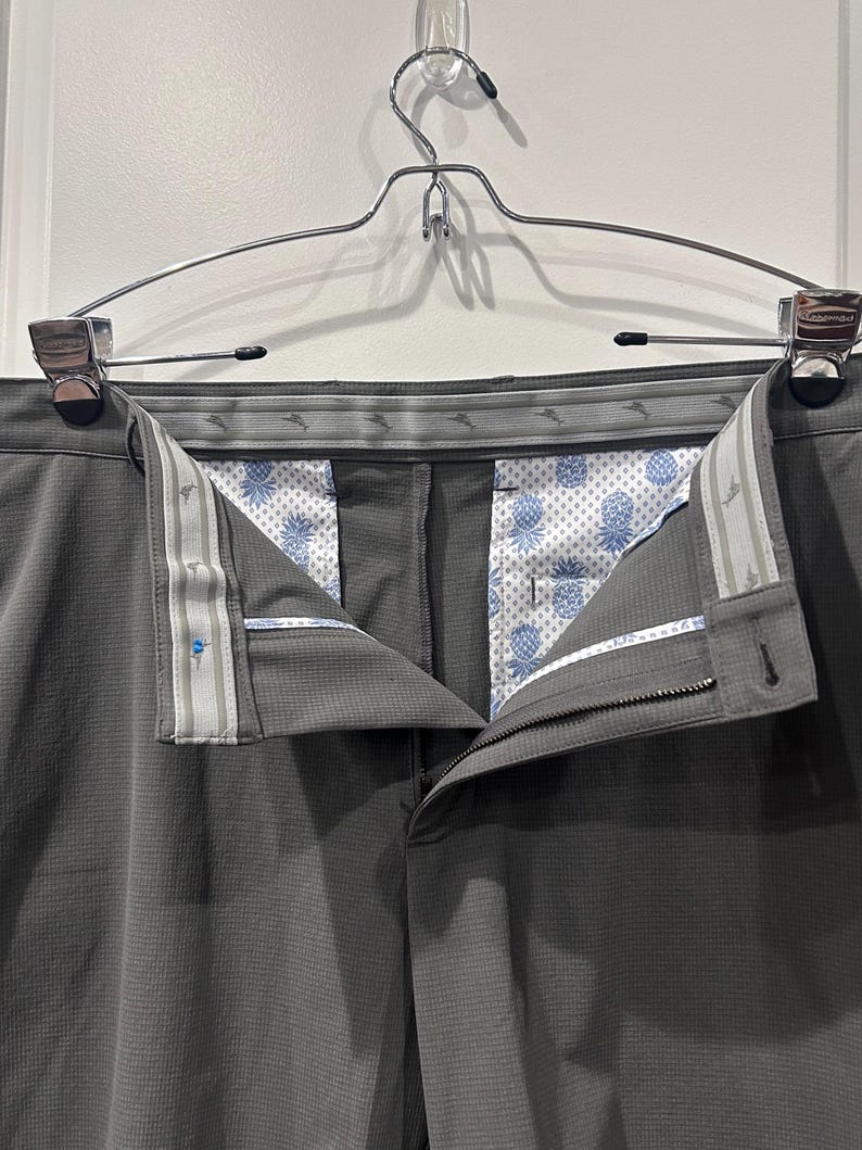 May include: Gray dress pants hanging on a silver hanger. The pants have a textured pattern and a button and zipper closure. The interior lining features a white background with a blue pineapple pattern. The waistband has a patterned design.