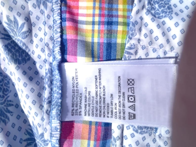 K&ouml;nnte beinhalten: Nahaufnahme des Hemdeninneren mit einem Etikett, das die Stoffzusammensetzung angibt: 62 % recyceltes Nylon, 33 % recyceltes Polyester und 5 % Elasthan. Waschhinweise sind ebenfalls vorhanden. Das Hemd hat ein blau-wei&szlig;es Ananasmuster.