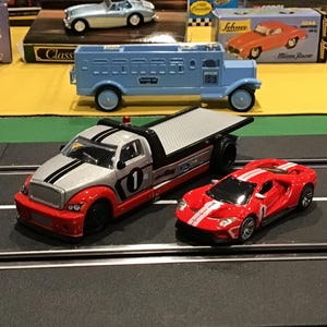 May include: A silver and red toy tow truck with a flatbed and a red race car with white stripes on a black track.