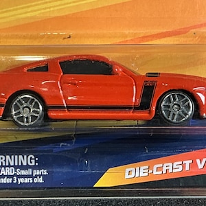 May include: A bright orange die-cast toy car, specifically a Ford Mustang Boss 302 model, featuring black side stripes and black hood accents. The vehicle has silver-colored wheels and is encased in its original clear plastic retail packaging.