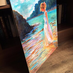 May include: An original oil painting on canvas depicts a person in a white dress standing on a beach. The artwork features a serene coastal scene with a blue sky, turquoise water, and colorful reflections. The person has light brown hair and a pink flower.