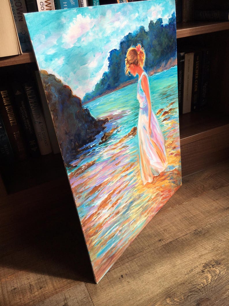 May include: An original oil painting on canvas depicts a person in a white dress standing on a beach. The artwork features a serene coastal scene with a blue sky, turquoise water, and colorful reflections. The person has light brown hair and a pink flower.