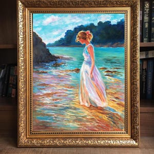 May include: An oil painting in a gold ornate frame depicts a woman in a white dress standing in shallow water. The artwork showcases a coastal scene with a blue sky, turquoise water, and colorful reflections. The frame is a detailed, gold-colored design.