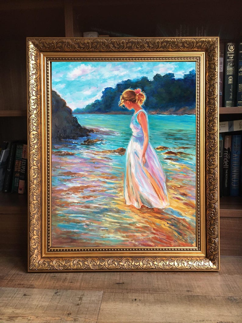 May include: An oil painting in a gold ornate frame depicts a woman in a white dress standing in shallow water. The artwork showcases a coastal scene with a blue sky, turquoise water, and colorful reflections. The frame is a detailed, gold-colored design.
