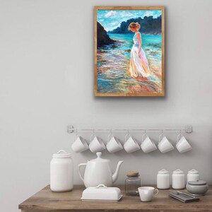 May include: A framed painting depicts a person in a white dress standing in shallow water. The artwork features a coastal scene with turquoise water, a rocky shoreline, and a blue sky. The frame is a light brown color.