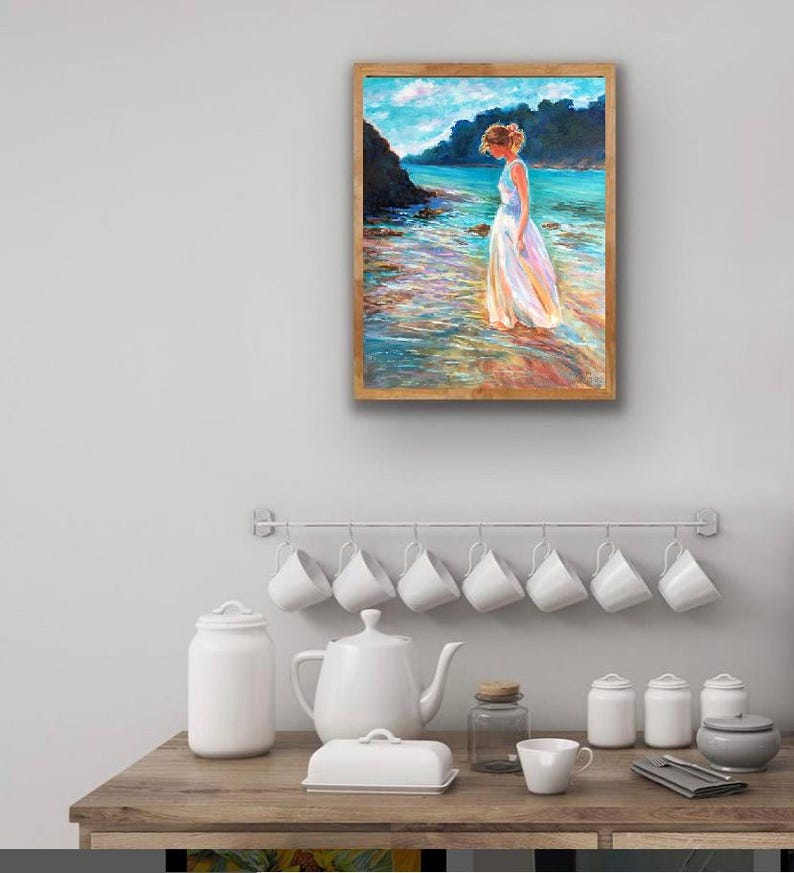 May include: A framed painting depicts a person in a white dress standing in shallow water. The artwork features a coastal scene with turquoise water, a rocky shoreline, and a blue sky. The frame is a light brown color.