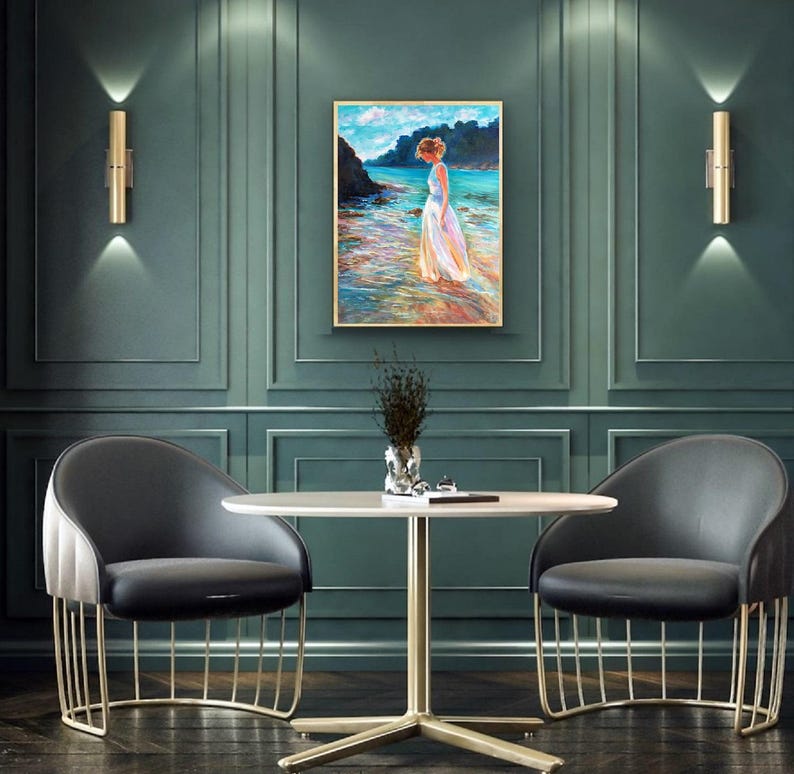 May include: An interior design scene featuring a painting of a woman in a white dress standing in the ocean. The painting is framed and hangs on a dark teal wall. Two gold sconces flank the painting. Two black chairs with gold frames and a round table complete the scene.