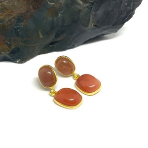 Puede incluir: Un par de pendientes de oro con dos piedras de cornalina en forma de lágrima. La piedra superior es de un tono naranja más claro, mientras que la piedra inferior es de un tono naranja más oscuro.