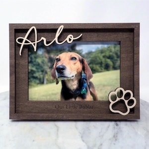 May include: A dark brown wooden picture frame featuring the name Arlo in script lettering at the top left and a paw print cutout at the bottom right. The frame includes the engraved text Our Little Bubba at the bottom centre.