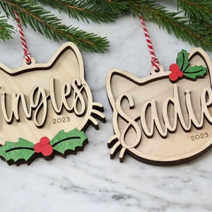 May include: Two wooden cat-shaped Christmas decorations with the names "Jingles" and "Sadie" and the year "2023" inscribed on them. Each decoration has a green holly sprig with red berries.