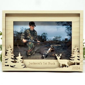 May include: A wooden picture frame with a cut-out design of trees and a deer. The frame features a photo of a young person holding a rifle in a wooded area. The text "Jackson's 1st Buck" and "2024" are engraved on the frame.