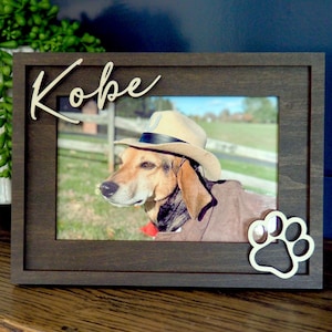 May include: A dark brown wooden picture frame featuring the name Kobe in white script lettering at the top left and a white paw print cutout at the bottom right. The frame holds a photograph of a dog wearing a tan hat.