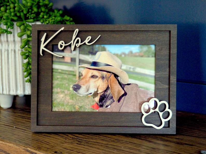 May include: A dark brown wooden picture frame featuring the name Kobe in white script lettering at the top left and a white paw print cutout at the bottom right. The frame holds a photograph of a dog wearing a tan hat.