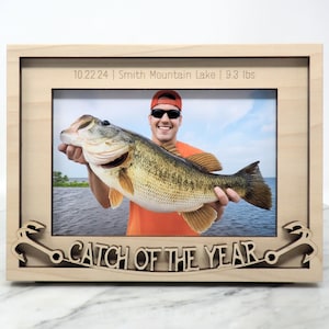 May include: Wooden picture frame with a photo of a person holding a large fish. The frame has the text "10.22.24 | Smith Mountain Lake | 9.3 lbs" and "CATCH OF THE YEAR". The frame is made of light-colored wood.