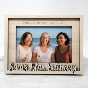 May include: A wooden photo frame displaying a picture of three women on a beach. The frame is inscribed with "Sara's 50th Birthday" and "Ocean City, Maryland | June 19, 2024". The women are smiling and holding glasses.