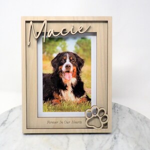 May include: A light-coloured wooden picture frame featuring the name Macie in script lettering at the top and the text Forever In Our Hearts at the bottom. A cutout paw print design is attached to the lower right corner of the frame.