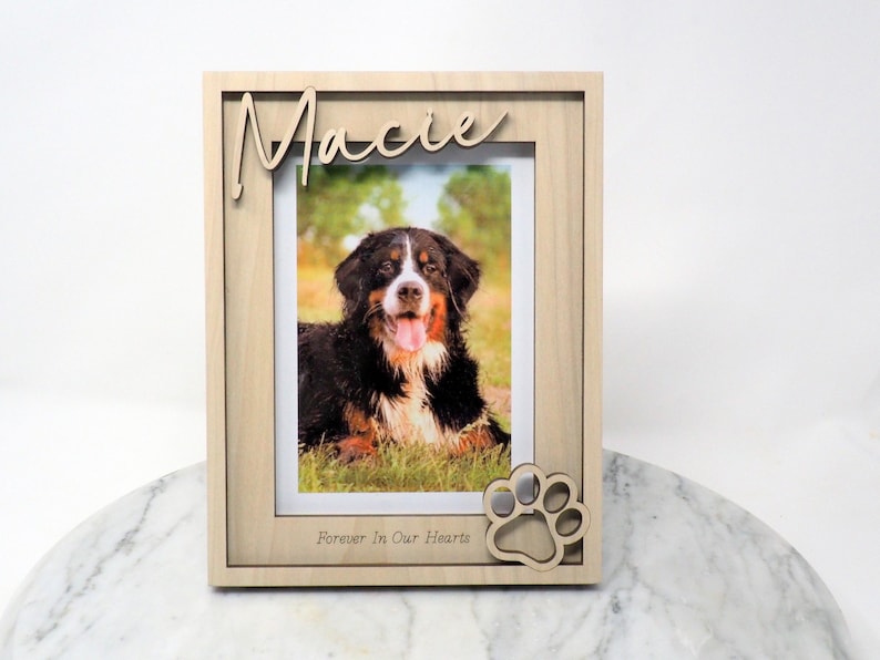 May include: A light-coloured wooden picture frame featuring the name Macie in script lettering at the top and the text Forever In Our Hearts at the bottom. A cutout paw print design is attached to the lower right corner of the frame.
