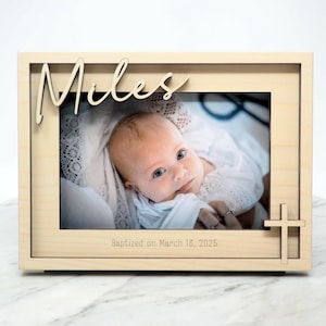 May include: A light-colored wooden picture frame with the name "Miles" in cursive script. The frame holds a photo of a baby and has a cross design on the lower right. The text "Baptized on March 16, 2025" is printed at the bottom.