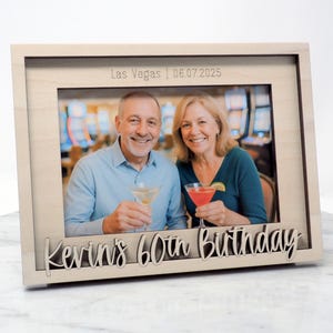 May include: A light-colored wooden picture frame with a photo of a smiling couple holding cocktails. The frame has the text "Las Vegas | 06.07.2025" and "Kevin's 60th Birthday" in a decorative font.