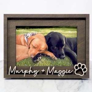 May include: A wooden picture frame with a photo of two dogs, one brown and one black, lying on grass. The frame has the names "Murphy + Maggie" and a paw print cutout.
