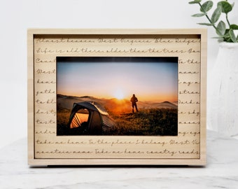 May include: A wooden picture frame with a landscape photo of a person standing near a tent at sunset. The frame has etched text around the photo. The text includes the words "Almost heavens West Virginia Blue Ridge".