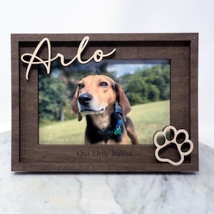 May include: A dark brown wooden picture frame featuring the name Arlo in script lettering at the top left and a paw print cutout at the bottom right. The frame includes the engraved text Our Little Bubba at the bottom center.