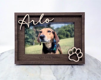 May include: A dark brown wooden picture frame featuring the name Arlo in script lettering at the top left and a paw print cutout at the bottom right. The frame includes the engraved text Our Little Bubba at the bottom center.