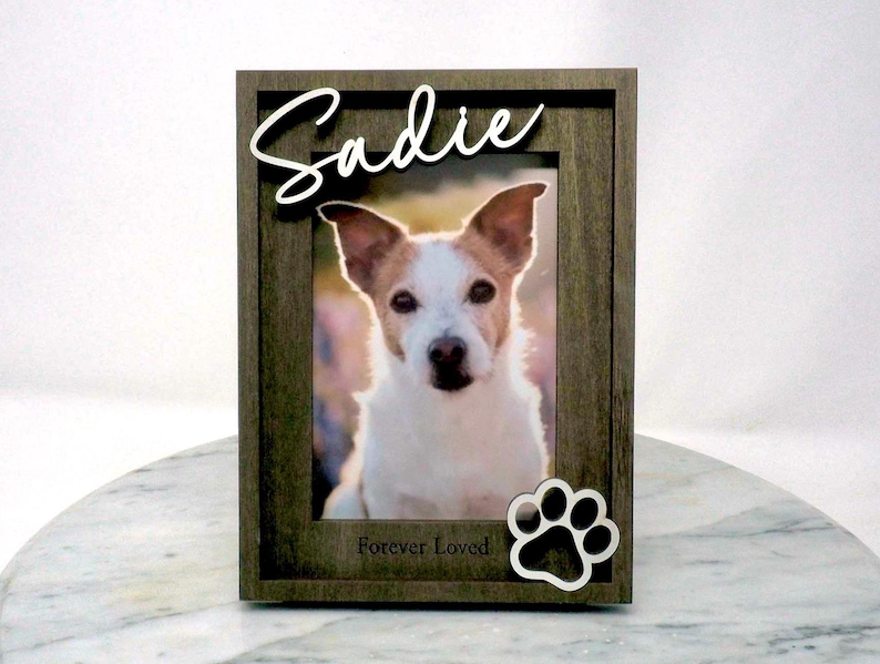 May include: A rectangular dark grey wooden picture frame containing a photograph of a white and brown dog. The name Sadie is written in white script at the top, and the words Forever Loved are printed below with a white paw print icon.