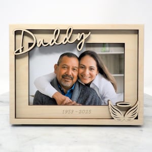 May include: Wooden photo frame with the word "Daddy" in script. The frame holds a photo of a man and woman. The dates "1953-2025" are engraved at the bottom. A decorative angel wing design is on the right side.