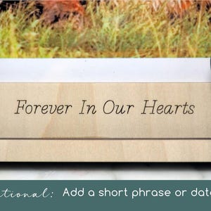 May include: A light-coloured wooden picture frame featuring the engraved text "Forever In Our Hearts" on the bottom border, with a cutout paw print design on the right side and a photograph visible in the upper section of the frame.
