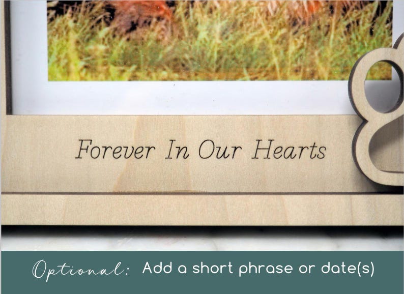 May include: A light-coloured wooden picture frame featuring the engraved text "Forever In Our Hearts" on the bottom border, with a cutout paw print design on the right side and a photograph visible in the upper section of the frame.