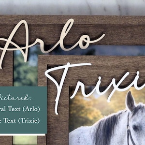May include: A dark brown wooden picture frame featuring the name Arlo in natural wood lettering and the name Trixie in white lettering. A photograph of a white horse is displayed within the frame. A teal box contains the text Pictured: Natural Text (Arlo) White Text (Trixie).
