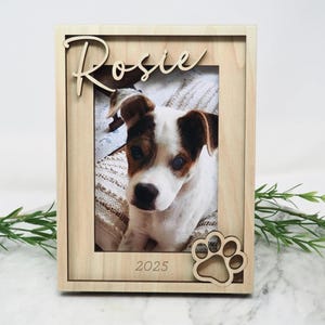 May include: A light-coloured wooden picture frame featuring the name Rosie in cursive script at the top, the year 2025 engraved at the bottom, and a cutout paw print design in the corner, containing a photograph of a white and brown puppy.