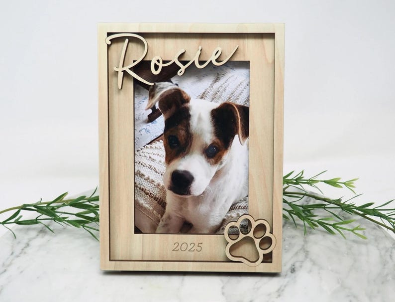 May include: A light-coloured wooden picture frame featuring the name Rosie in cursive script at the top, the year 2025 engraved at the bottom, and a cutout paw print design in the corner, containing a photograph of a white and brown puppy.