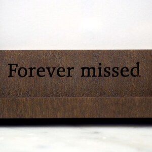 May include: A dark brown wooden frame featuring the engraved text Forever missed in a serif font. The frame has a smooth, flat horizontal base and a portion of a light-coloured wooden letter visible on the right side of the structure.