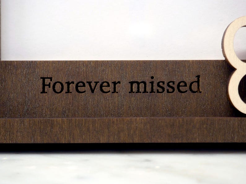 May include: A dark brown wooden frame featuring the engraved text Forever missed in a serif font. The frame has a smooth, flat horizontal base and a portion of a light-coloured wooden letter visible on the right side of the structure.