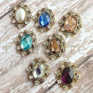 May include: Assortment of eight oval-shaped, decorative embellishments with rhinestones and various colored glass gems. The embellishments are in shades of blue, purple, pink, brown, and white.