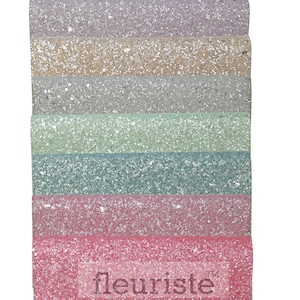 May include: A stack of six glitter fabric sheets in various pastel colors, including light purple, beige, light gray, light green, teal, and pink. The fabric sheets are stacked on top of each other and have a glittery texture. The bottom sheet has the word "fleuriste" printed on it.