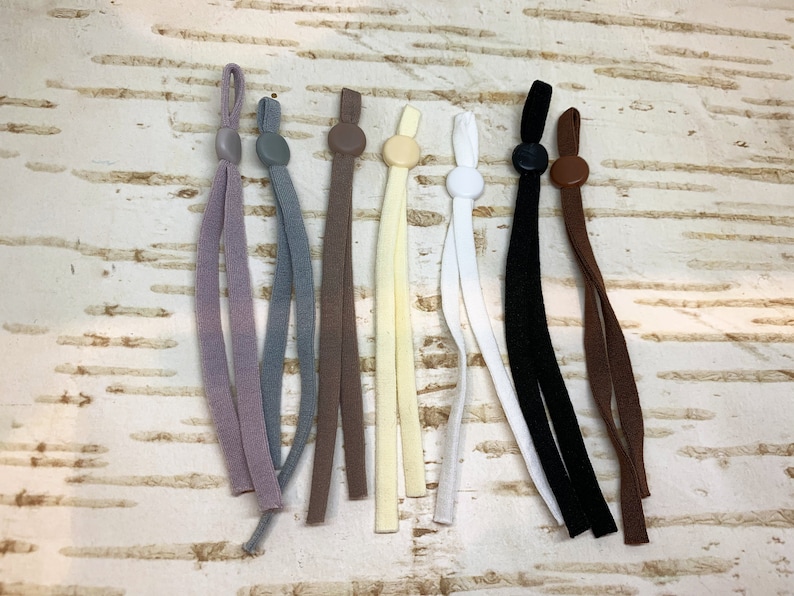 K&ouml;nnte beinhalten: Sortiment von acht elastischen Schnurextendern f&uuml;r Masken mit verschiedenen farbigen Schn&uuml;ren und Plastikstopfen. Die Schn&uuml;re sind in den Farben Braun, Grau, Beige, Wei&szlig;, Schwarz und Lila.